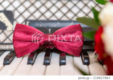 A Maroon Bow Tie Resting on Piano Keys With Floral Arrangements Nearby. 119827863