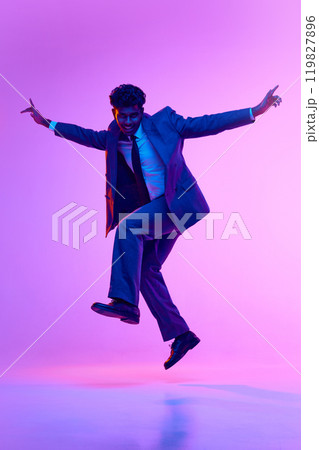 Full-length portrait of happy young Indian man, businessman in formal wear dancing, jumping in joy against purple studio background in neon light 119827896