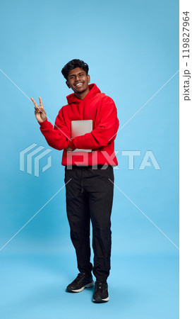 Full-length. Young Indian man in casual clothes using tablet for online communication, standing with funny meme face against blue studio background 119827964