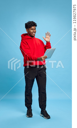 Full-length image of young Indian man in casual clothes holding tablet, having online video call, waving in greeting against blue studio background 119827975