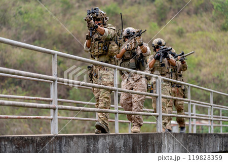 Group of militaries or soldiers with battle suit hold and point gun and walk in the row on bridge during outdoor practice. 119828359