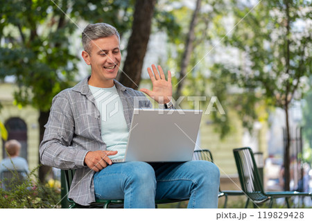 Mature man sitting in city street looking at laptop camera making video webcam conference call Mature man sitting in city street looking at laptop camera making video webcam conference call 119829428