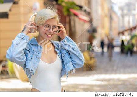 Portrait of smiling young woman in glasses taking off wireless headphones while looking at camera Portrait of smiling young woman in glasses taking off wireless headphones while looking at camera 119829446