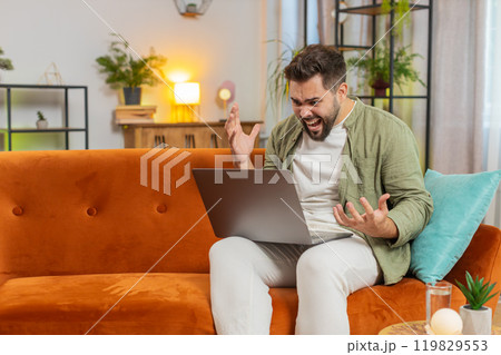 Upset young Caucasian man freelancer using laptop engages in heated conversations sitting on sofa 119829553