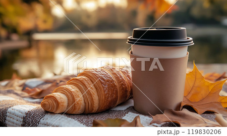 Coffee or tea cup and croissant on picnic plaid near the lake in autumn park. Cozy autumn picnic set up outdoors in the morning, breakfast in nature 119830206