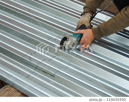 A man in work clothes and a glove uses an angle grinder to cut or carve a metal sheet or roofing material along markings 119830300