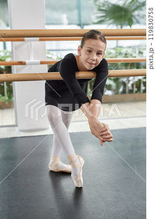 Vertical shot of young ballerina in pointe shoes cheerfully smiling posing for camera while leaning on barre in dance studio 119830758