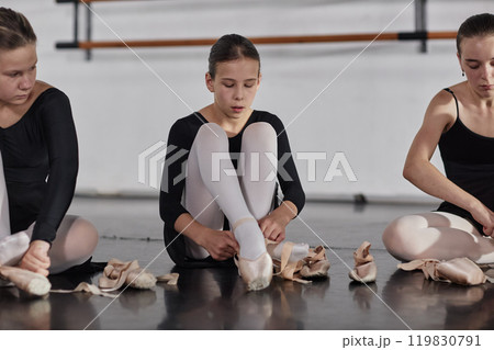 Concentrated young girl putting on pointe shoes preparing for ballet class while sitting on floor with kids in dance studio, copy space Concentrated young girl putting on pointe shoes preparing for ballet class while sitting on floor with kids in dance studio, copy space 119830791