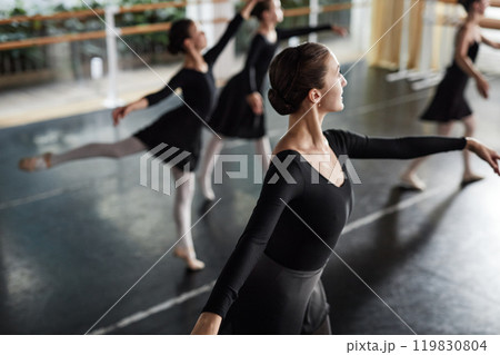Graceful female dance teacher dressed in black leotard supporting little concentrated girl with hands while kid standing on one leg learning ballet positions during class in studio, copy space Graceful female dance teacher dressed in black leotard supporting little concentrated girl with hands while kid standing on one leg learning ballet positions during class in studio, copy space 119830804