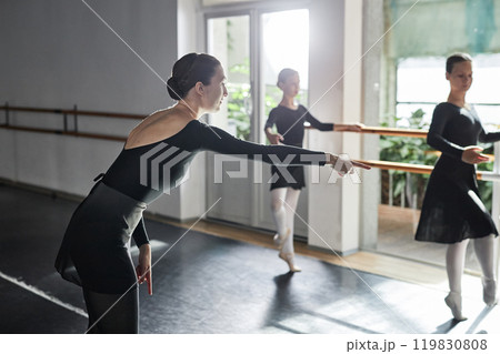 Side view of female choreographer in leotard with back cut pointing with finger drawing attention to leg positioning while teaching kids ballet moves during group class in dance studio, copy space 119830808