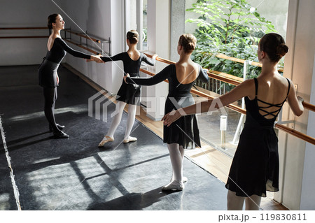 Rear view of three young girls wearing elegant dance leotards learning ballet positions at barre while female choreographer assisting children in sunlit studio 119830811