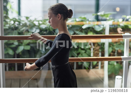 Side view of concentrated teenage girl in elegant black leotard practicing ballet dance positions standing at barre during class in studio, copy space Side view of concentrated teenage girl in elegant black leotard practicing ballet dance positions standing at barre during class in studio, copy space 119830818