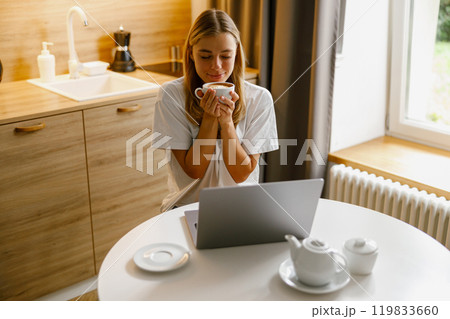 A Woman is Enjoying a Cup of Coffee While Working on Her Laptop in a Modern Stylish Kitchen 119833660