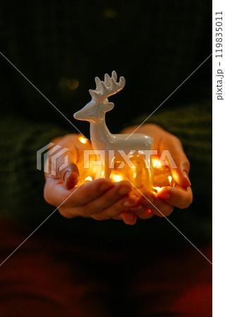 Close-up of woman hands with illuminated Christmas deer figurine. Woman hands gently hold a ceramic deer figurine illuminated with warm light, Christmas spirit and festive warmth of winter season 119835011