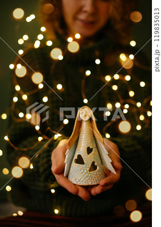 Close-up of hands holding angel figurine with Christmas bokeh background. Woman hands gently holding glowing ceramic angel, concept Christmas and Angel Day spirit in cozy atmosphere with bokeh lights Close-up of hands holding angel figurine with Christmas bokeh background. Woman hands gently holding glowing ceramic angel, concept Christmas and Angel Day spirit in cozy atmosphere with bokeh lights 119835013