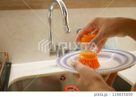 Hands scrub a plate with an orange brush in a sink, emphasizing cleanliness and hygiene 119835850