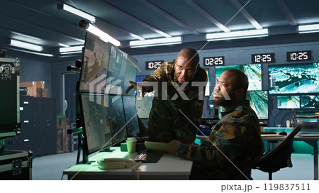 Military officers look at intel folder files on enemy battle formation, comparing with radar images. Army specialists looking at mission case file and equipment screens, camera B 119837511