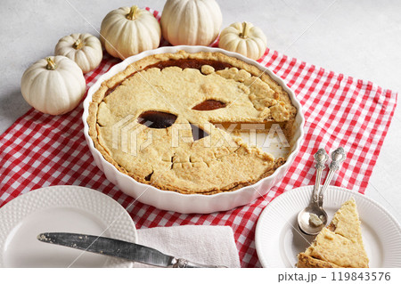 Halloween food. Homemade pumpkin pie or tart with a scary face for Halloween on a red napkin on gray table with cut piece on the plate. Copy space. Halloween food concept. 119843576