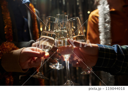 Cropped shot of friends clinking glasses of champagne standing in top light while celebrating Christmas in living room with silver tinsel at home, copy space Cropped shot of friends clinking glasses of champagne standing in top light while celebrating Christmas in living room with silver tinsel at home, copy space 119844018