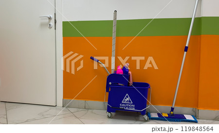 Bucket trolley with detergents and mop for cleaning floor against background of wall and door in corridor, cleanliness in public place, cart on wheels for cleaning service employee, caution wet floor Bucket trolley with detergents and mop for cleaning floor against background of wall and door in corridor, cleanliness in public place, cart on wheels for cleaning service employee, caution wet floor 119848564