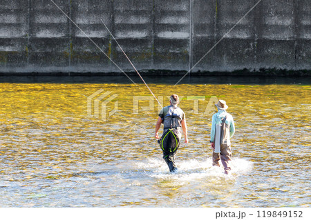 Two fishermen wading in shallow river waters near a concrete dam, ready for sturgeon fishing, serene scene blending nature and industrial backdrop Two fishermen wading in shallow river waters near a concrete dam, ready for sturgeon fishing, serene scene blending nature and industrial backdrop 119849152