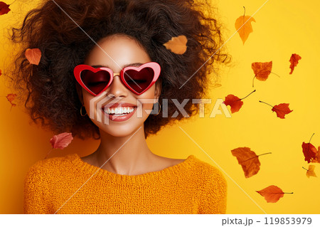 Happy young woman wearing heart shaped sunglasses surrounded by fallen autumn leaves. Autumn love, romance fall. Autumn fashion, lifestyle and holidays. Hello autumn concept 119853979