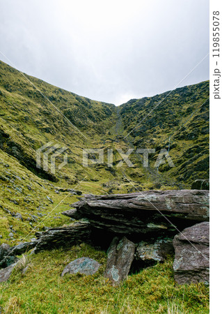 A serene valley with vibrant greenery, flanked by rugged slopes under a cloudy sky. A large rock sits prominently in the foreground, enhancing the natural beauty. 119855078