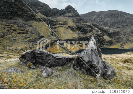 A striking rocky outcrop is prominently featured in the foreground, while a waterfall cascades down the mountainside into a tranquil lake surrounded by lush greenery. 119855079