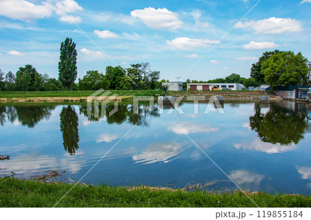 The river flows through the city. On the calm surface of the river banks are reflected. Slovak river Nitra on a bright sunny day. The river flows through the city. On the calm surface of the river banks are reflected. Slovak river Nitra on a bright sunny day. 119855184