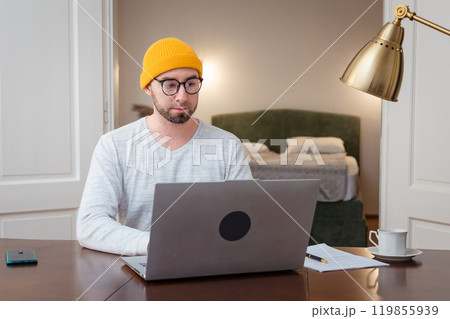 Caucasian adult man wearing hat and glasses is working at laptop. Remote work from the home office. The student is studying remotely. Concept of freelancing Caucasian adult man wearing hat and glasses is working at laptop. Remote work from the home office. The student is studying remotely. Concept of freelancing 119855939