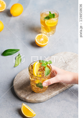 A hand takes a glass of iced tea with lemon and mint against a gray background with citrus fruits. 119858636