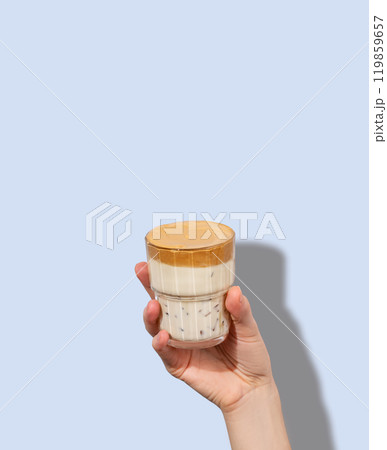 Dalgona coffee. A hand holds a glass of whipped and instant iced coffee and milk in a glass  119859657