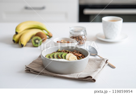 Baked granola with banana and kiwi on the table with cup of tea on a white kitchen background. Baked granola with banana and kiwi on the table with cup of tea on a white kitchen background. 119859954