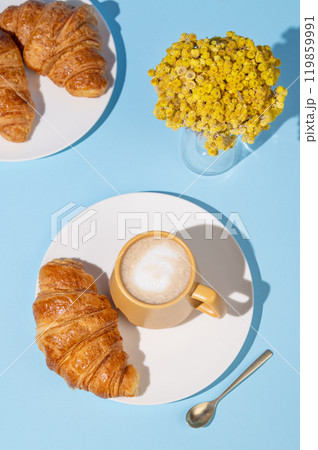 Flat lay of coffee cup and fresh croissants on blue background with yellow flowers and shadow. Flat lay of coffee cup and fresh croissants on blue background with yellow flowers and shadow. 119859991