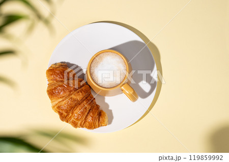 Flat lay of coffee cup and fresh croissant on a white plate on a yellow background  119859992