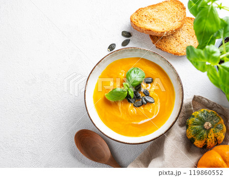 Flat lay of pumpkin soup with cream, basil and seeds on a light background with bread, spoon 119860552
