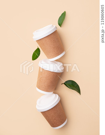 Three brown paper coffee cups with white lids on beige background with green leaves. Three brown paper coffee cups with white lids on beige background with green leaves. 119860605