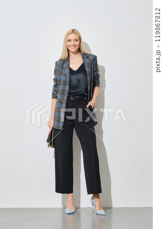 Cheerful woman in stylish checkered jacket and wide trousers posing by white wall 119867812