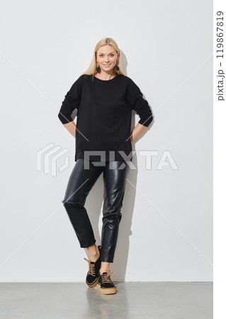 Smiling woman in pullover and leather trousers leaning against white wall 119867819