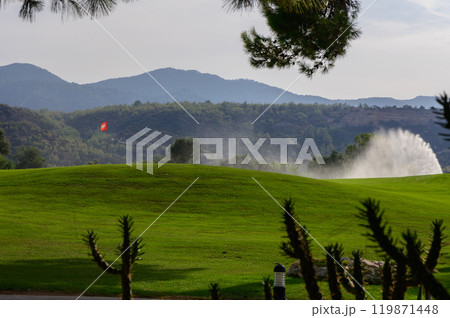 Serene golf course landscape with misty fountain and distant mountains under bright sunlight in a tranquil afternoon setting 119871448