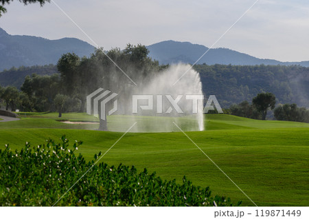 A tranquil golf course backdrop featuring a lively sprinkler amidst lush green fairways and distant mountains under a bright sky 119871449