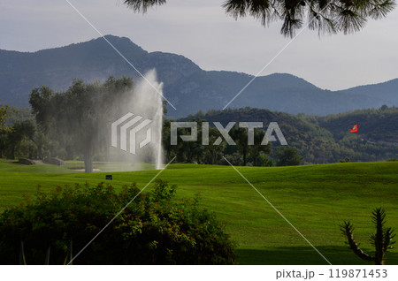 A serene landscape featuring a golf course with a water fountain amidst rolling hills under a clear sky in the early morning light 119871453