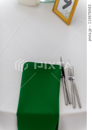 Close up of wine glasses, silver tableware and green napkins on round tables covered with white tablecloth. 119876083