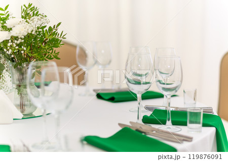 Close up of wine glasses, silver tableware and green napkins on round tables covered with white tablecloth. 119876091