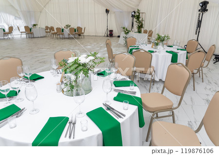 Wine glasses, silver tableware and green napkins on round tables covered with white tablecloth. 119876106
