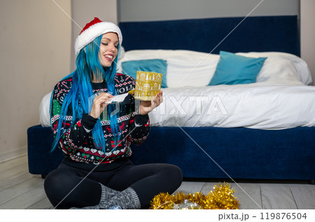Portrait of a beautiful caucasian hipster woman in Christmas clothes. She is sitting next to the bed and smilingly unwrapping a package. Expectation, surprise, happiness. Portrait of a beautiful caucasian hipster woman in Christmas clothes. She is sitting next to the bed and smilingly unwrapping a package. Expectation, surprise, happiness. 119876504