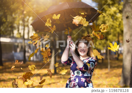 Beautiful young woman in an autumn park with an umbrella from which yellow leaves are falling. 119878128