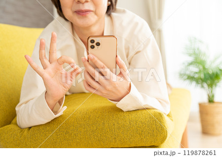ソファに寝そべってスマホを見ながら丸サインをするミドル〜シニア女性 119878261