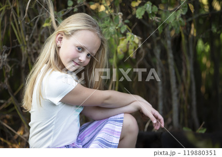 The little blonde girl sits near the green bushes and looks at the camera. 119880351
