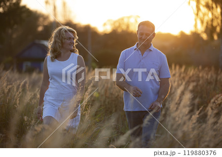 A beautiful middle-aged couple in love are walking on the field against the background of sunset. 119880376
